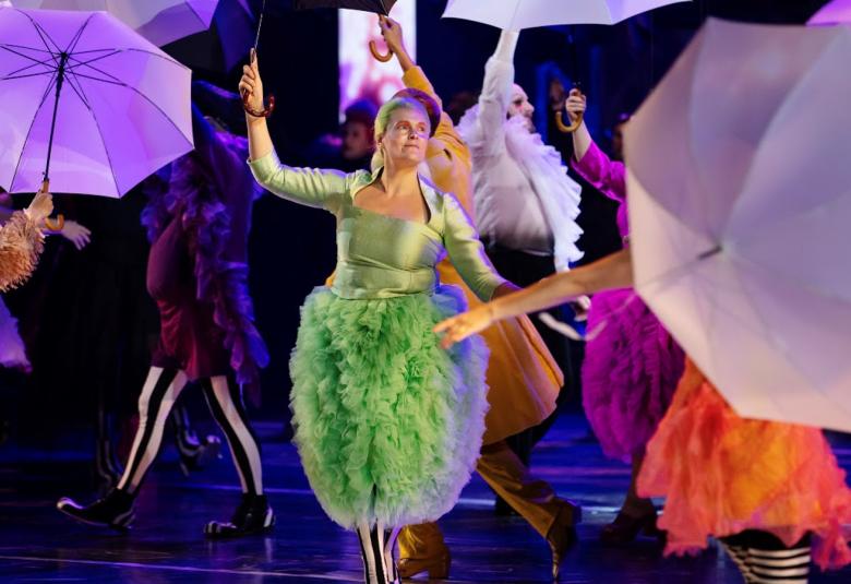 Kuvassa on värikkäästi pukeituneita, ympäriinsä käveleviä henkilöitä ja heillä on vaalean violetit sateenvarjot kädessään.   Edessä keskellä on nainen vaalean vihreässä mekossa, jonka alaosa on röyhelöinen.