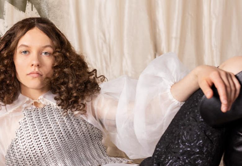 Kuvassa on Elsi Sloan istumassa nojaten vasempaan käteensä ja hänellä on hopeinen virkattu pusero ja läpinäkyva, valkoinen pusero sen päällä sekä mustat housut. Taustalla on vaaleaa kangasta.