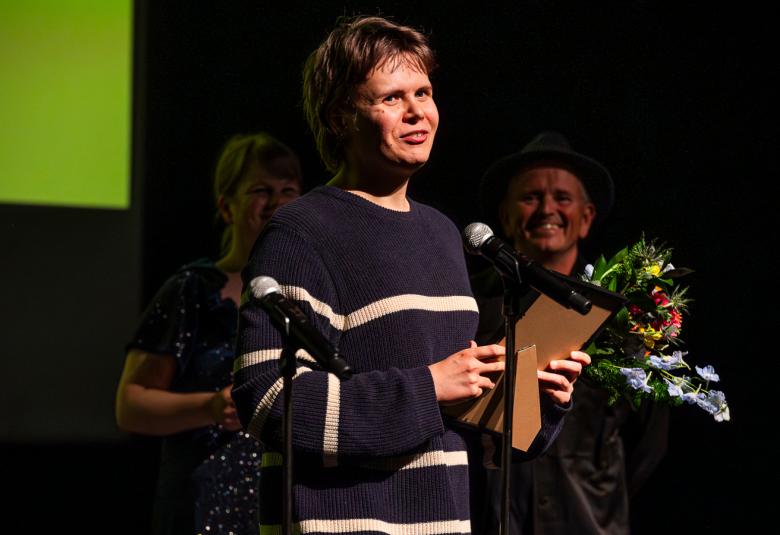 Kuvassa on keskellä valkoisella isoin krijaimin Sebastian. Taustalla on miehen otsa ja hän on kumartuneena kohti edessä olevan nuoren miehen selkää. Nuoresta miehestä näkyy vain puolet kasvoista.