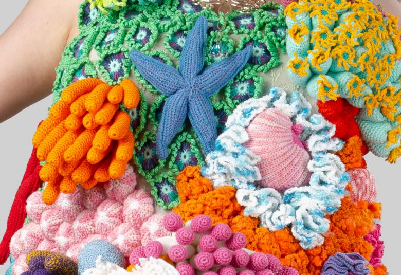 Kuvassa on mekko, jossa on virkattuna vihreitä ruutuja, porkkanoita, vaalean punaisia retiisejä ja eri värisä kohokuvioita. Käsivarsia näkyy osittain.