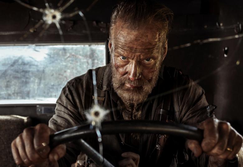 Kuvassa on Jorma Tommila ajamassa autoa kädet ratissa. Tuulilasissa on kadessa kohtaa luodin reiät.  Takana näkyy auton ikkuna. Kuvan sävy on tumma.