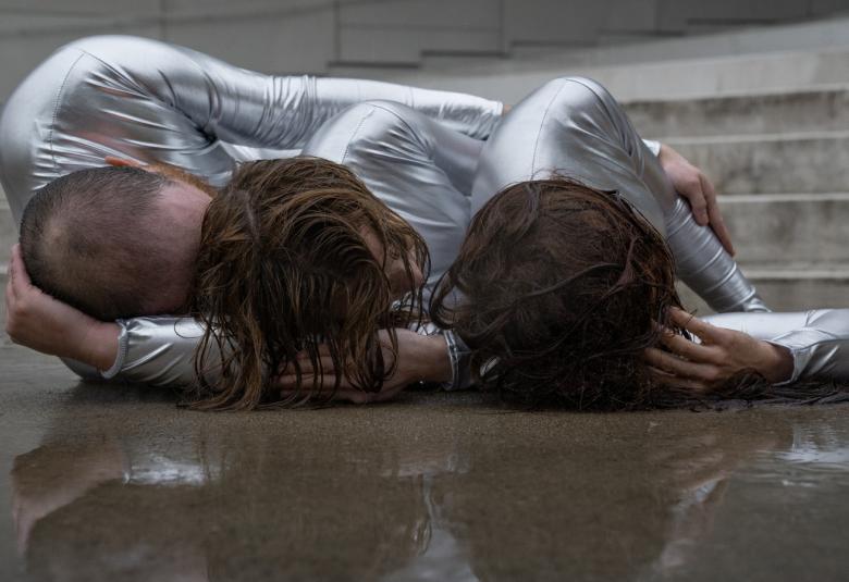 Kuvassa on kolme tanssijaa lattialla kuvattuna pääpuolesta käpertyneenä lattialle. Kuva on musta-valkoinen.