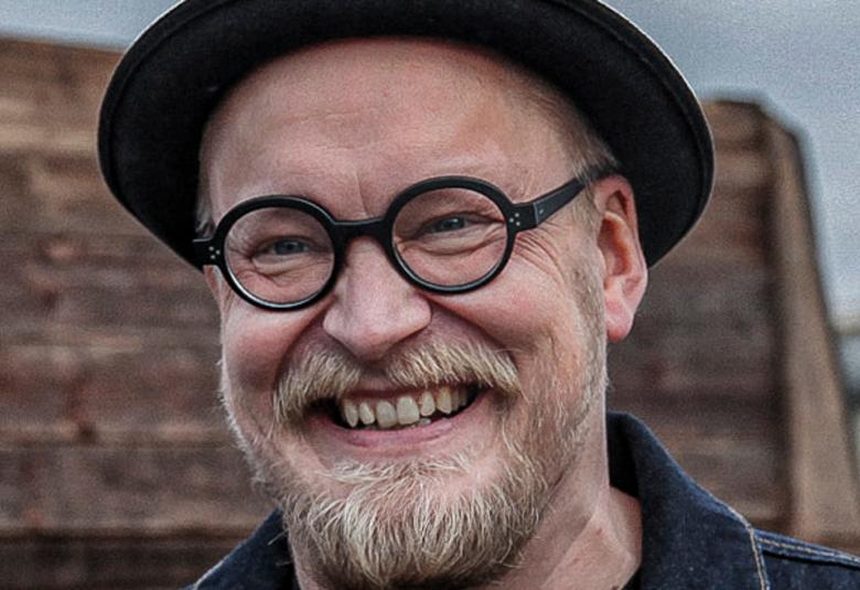 Kuvassa on Teemu Nikki kasvokuvassa. Hänella on tumma hattu päässään ja vaaleaa partaa. Taustalla on ruskeaa rakennusta.