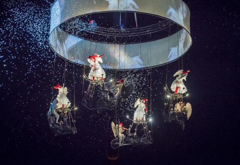 Kuvassa on kuusi sirkustaiteilijaa vaaleat mekot päällään roikkumassa isosta metallisesta lieriöstä metallisissa kulkuneuvoissa.  Taustalla on tummalla taustalla valopilkkuja.