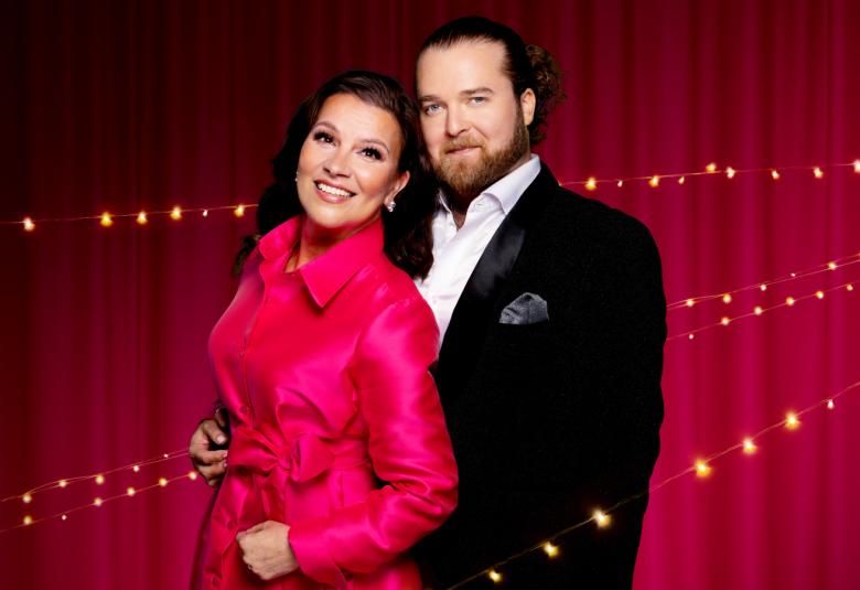 Kuvassa ovat ARja Koriseva vasemmalla punaisessa puvussa. Oikealla on Teemu Roivainen mustassa takissa ja valkoisessa paidassa. He ovat puolivartalokuvassa. Tausta on tumman punainen.