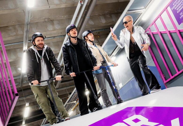 Kuvassa on alhaalta ylöspäin kuvattuna neljä elokuvan miesnäyttelijää tummiss tai vaaleissa pusakoissa ja tummissa housuissa. Sivustoissa on violettia.