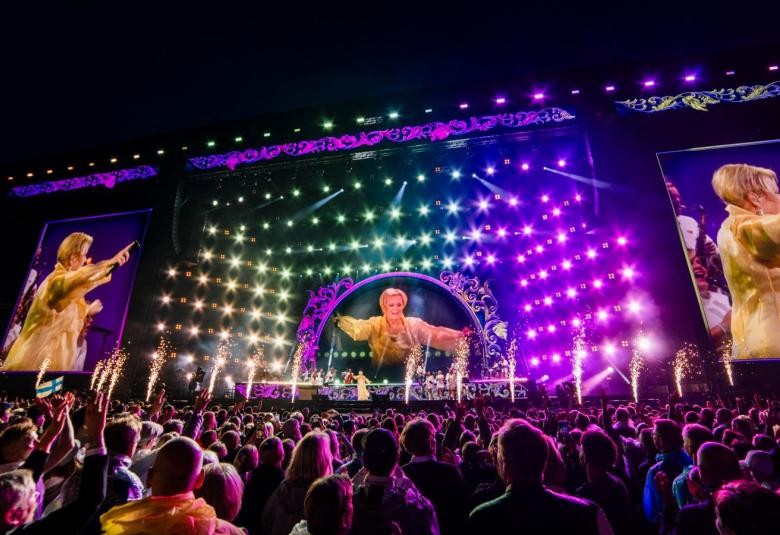 Kuvassa on näkymä, jossa ova vasemmalla ja oikealla näytöt reunoilla niissä Katri Helena on kultaisessa puvussa. Hän on sivuttain. Keskellä on iso estradi ja siinä valoja katossa sekä sivuilla. Takana on iso näyttö ja Katri Helena edestä päin. Edessä on yleisöä selin. Kuvassa on pimeää.