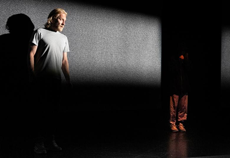 Kuvassa Tuukka Huttunen seisomassa harmaassa t-paidassa ja nurkassa on Milla Tissari kyykyssä. Puolet kuvasta on tummaa.