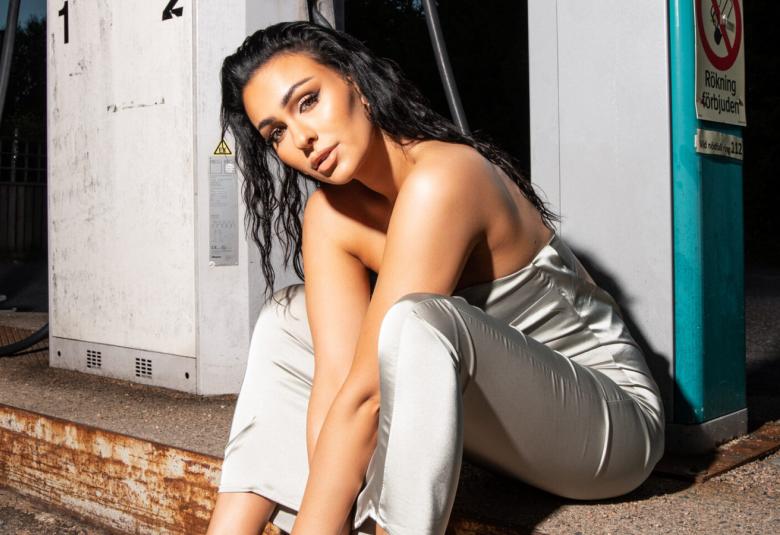 Kuvassa on Sara Siipola istumassa portaalla ja takana on valkoiset pylväät. Hän nojaa eteenpäin kädet polvien välissä. Siipolalla on valkoinen housupuku,jossa ei ole hihoja ja olkapäät näkyvissä.