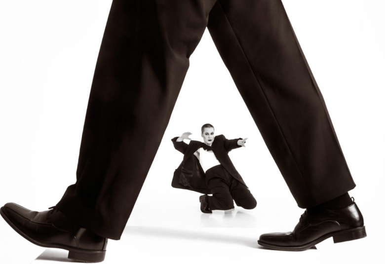 Kuvassa on isot jalat kävelemässä mustissa lahkeissa ja kengissä. Alta näkyy pienikokoisena miestanssija polvillaan mustassa puvussa ja valkoisessa paidassa. Tausta on valkoinen.