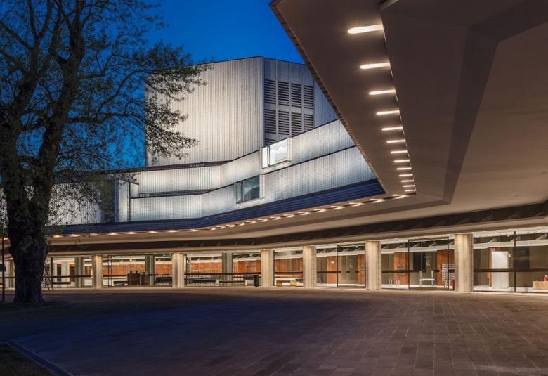 Kuvassa on iltakuva valaistuksessa olevasta Helsingin kaupunginteatterista. Alaosassa on ikkunarivistö ja yläosasassa kaksi kerrosta valkoista kattorakennelmaa ja päällä valkoinen torni. Vasemmalla on lehdetön puu.