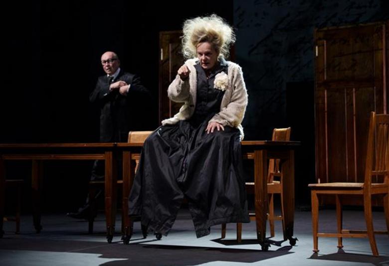 Kuvassa on Sari Puumalainen keskellä istumassa pöydän päällä mustassa, pitkässä mekossa ja vaaleassa villatakissa vaaleat hiukset pörrössä. Taustalla on Antti Pääkkönen seisomassa mustassa puvussa. Tausta on tumma.