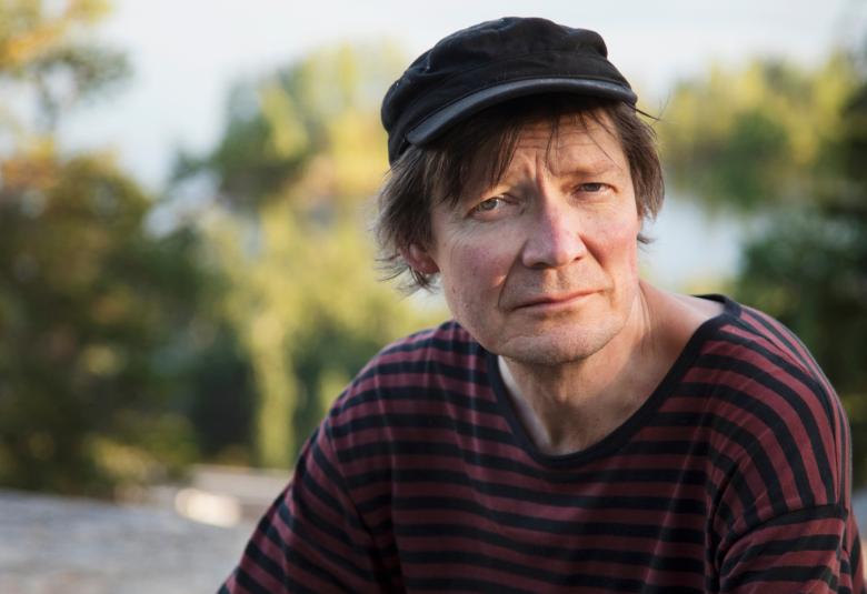 Kuvassa on Juha Hurme puolivartalokuvassa istumassa. Hänellä on violetti-mustaraitainen pusero päälään ja musta lippalakki päässään. Taustalla on kauempana näkyy vihreitä lehtipuita.