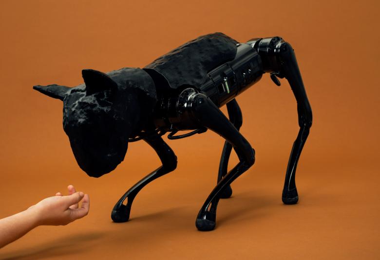 Kuvassa on musta metallinen robottikoira, joka on kumartuneena vasemmasta kulmasta ojentautuvaa kättä kohti. Tausta on oranssin ruskea.