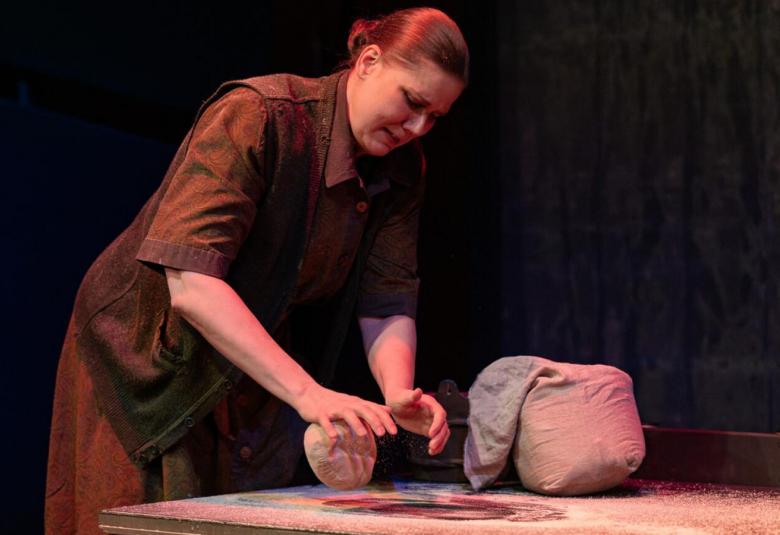 assa on nainen leopomassa ruisleipää hieman sivuttain. Hnellä on ruskea mekko päällään. Pöydällä vieressä pieni jauhosäkki.