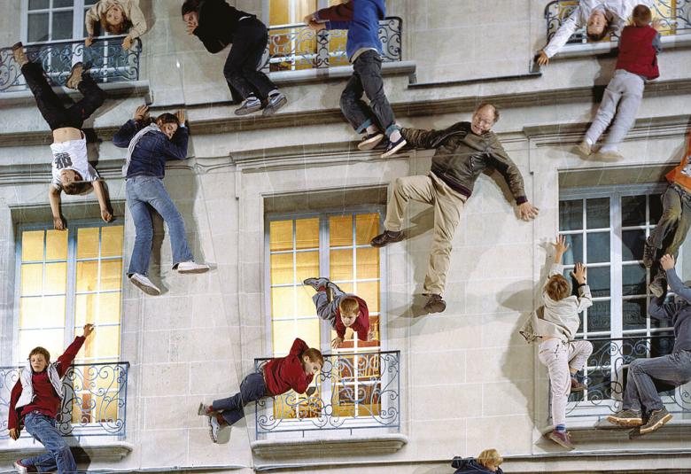 Kuvassa on ihmisten kokoisia nukkehahmoja roikkumassa harmaalla talon seinällä, jossa on runsaasti korkeita pikkuruutusia ikkunoita.