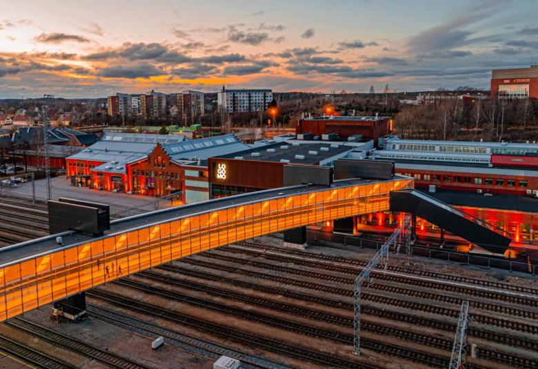 Kuvassa on Turun Logomon rakennukset ja sen ympäristöä iltahämärässä. Alaosassa on valaistu silta, joka on rautieraiteiden yläpuolella.
