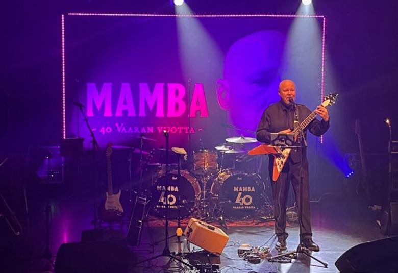 Kuvassa on keskellä Tero Vaara soittamassa kolmikulmaista kitaraa ja laulamassa mustassa paidassa ja pitkissä housuissa. Vieressä on bähdisoittimia. Taustalla on tumman violetilla valaistua lavaa ja seinällä teksti Mamba pinkillä.