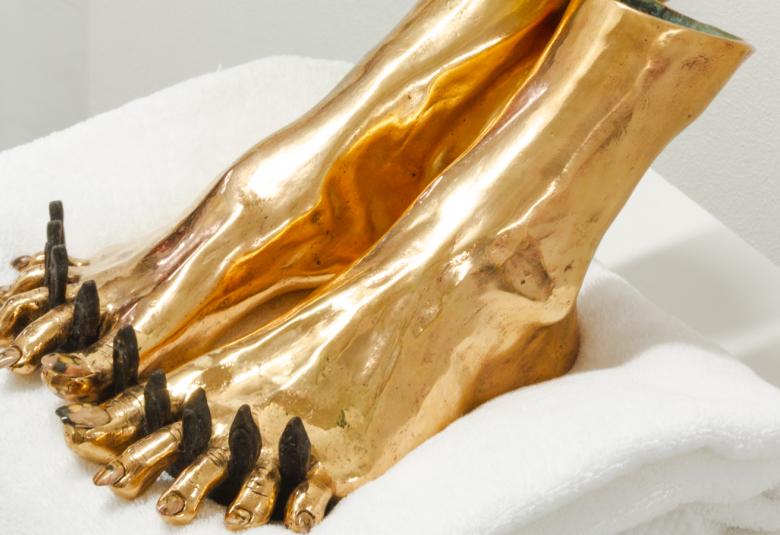 Kuvassa on kultaiset jalat jalkahoitotyynyllä ja niissä on mustat tupot varpaiden välissä. 