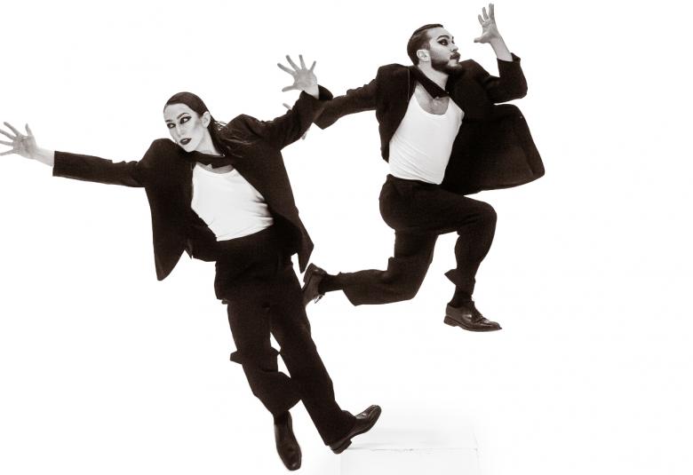 Kuvassa on vasemmalla naistanssija mustassa miesten puvussa kädet levitettyinä vinoasennossa. Oikealla on miestanssija myös mustassa puvussa ja valkoisessa paidassa juoksemassa. Tausta on valkoinen ja kuva on musta-valkoinen.