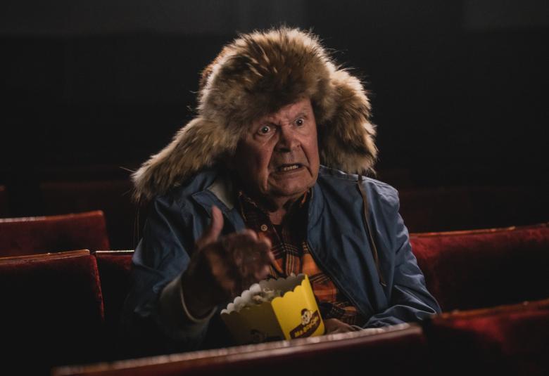 Kuvassa on Heikki Kinnunen Mielensäpahoittajana yksin istumassa elokuvateatterissa karvahattu päässään ja tummassa takissa. Tausta on tumma.