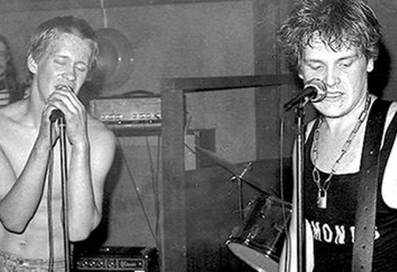 Kuvassa on vasemmalla Aku Syrjä laulamassa ilman paitaa ja oikealla on Mikko Saarela mustassa hihattomassa t-paidassa soittamassa kitaraa ja laulamassa mikrofoni edessään. Kuva on musta-valkoinen.