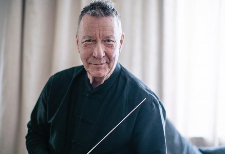 Kuvassa on Okko Kamu puolivartalokuvassa ja hänellä on tumma pusero päällään sekä tahtipuikko kädessään. Taustalla on vaaleata verhoa.