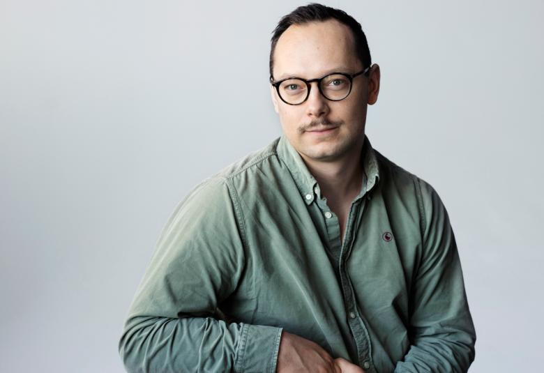 Kuvassa on Paavo Teittinen istumassa ja nojaa oikeaan kätensä. Hänellä on vihertävä paita. Tausta on harmaa.