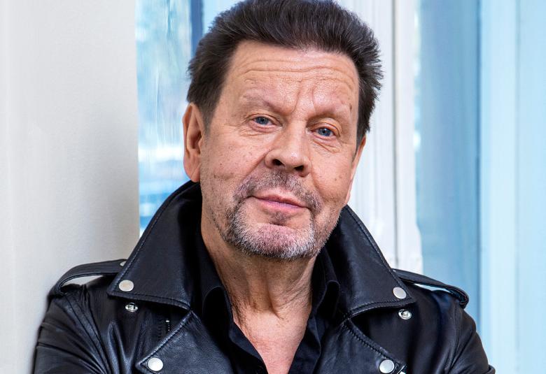 Kuvassa on Pate Mustajärvi puolivartalokuvassa.  Hänellä on musta nahkatakki päällään. Taustalla on vaaleata ja vaalean sinistä seinää.