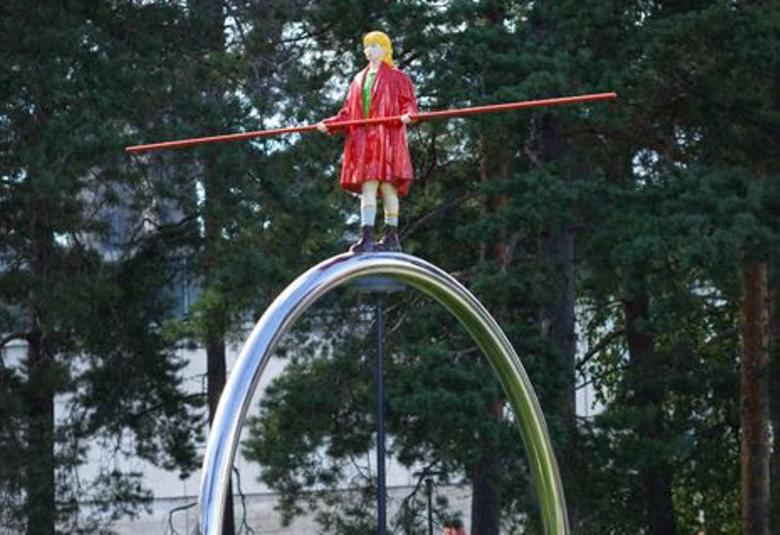 Kuvassa on vihreiden lehtipuiden edessä ison teräsrnkaan päällä punaisissa vaatteissa tasapainotteleva hahmo,jolla on piktä keppi vaakatasossa käsissään.