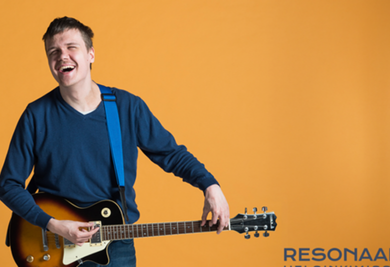 Kuvassa on nuori mies kitaraa soittamassa ja hänellä on sininen collegepaita päällään. Tausta on ruskehtavan keltainen. 