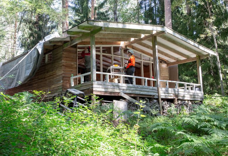 Kuvassa on vihreässä metsässä matala, loivasti harja kattoinen kesämökki, jossa on koko mökin pituinen terassa ja takan isot ikkunat. Terassilla seisoo mies.