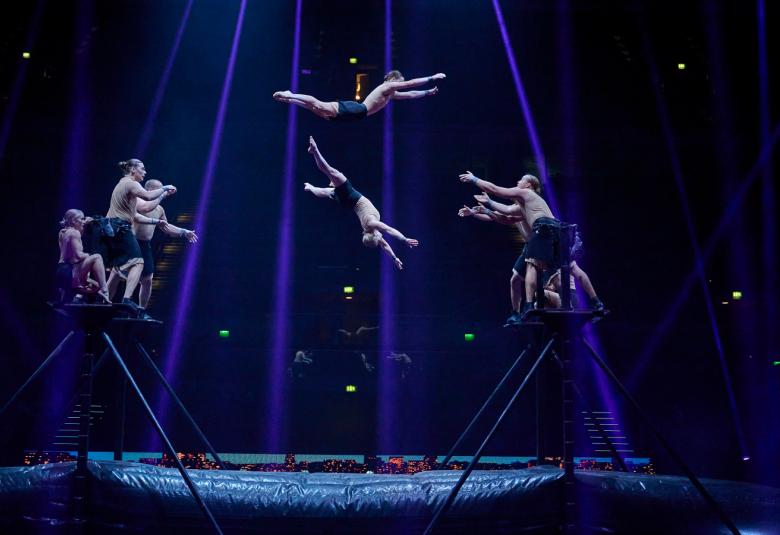 Kuvassa on sinisessä valokartiossa eri puolilla roikkuvassa sirkustaiteilijoita trapetsilla. Tausta on tumma.