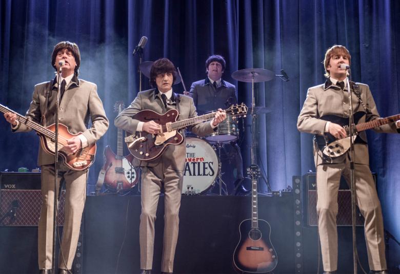 Kuvassa ovat The Beatles-tribuuttibändin kolme jäsentä edessä kitaroineen ja heillä on vaalean ruskeat puvut päällään. Taustalla on rumpali. Takana on tumman sininen verho.