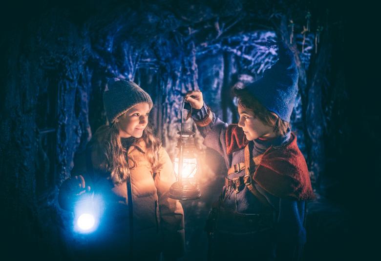 Kuvassa on tumman taustan keskellä valaistusta ja siinä on vasemmalla Soma Zimmatore pipo päässään ruskeassa toppatakissa ja lyhtyä pitelee  Marco Stylianou sinisessä pipossa. Taustalta näkyy sinistä luolaa.