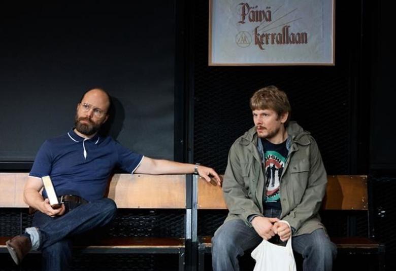 Kuvassa on vasemmalla istumassa penkillä sinisessä puserossa ja housuissa Juho Milonoff ja oikealla Paavo Kinnunen vihreässä maiharissa ja sinisissä farkuissa. Taustalla on tummaa seinää ja taulu, jossa lukee Päivä kerrallaan.