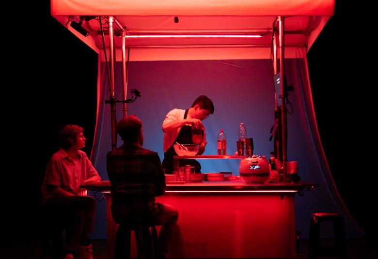 Kuvassa on Jaha Koo tekemässä ruokaa sivuttain telttakatoksellisessa keittiössä, joka on valaistu punaisella. Sivussa istuu baarituoleilla kaksi asiakasta tummana.