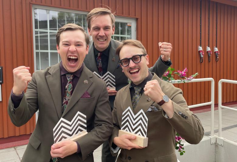 Kuvassa on KAJ:n kolme jäsentä ja he ovat ruskeissa puvuissa puolivartalokuvassa. Kaksi on edessä ja yksi takana. Heillä on musta-valkeasakarakuvioiset palkinnot kädessään. Taustalla on ruskea rakennus.