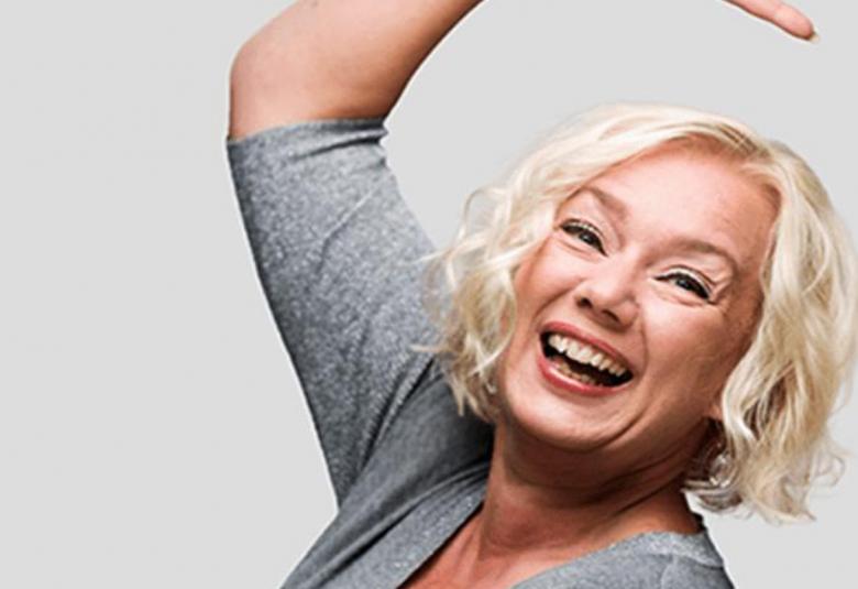 Kuvassa on Sari Siikander iloisesti oikea käsi ylös kohotettuna. Hänellä on harmaa pusero päällään ja vaaleat hiukset.