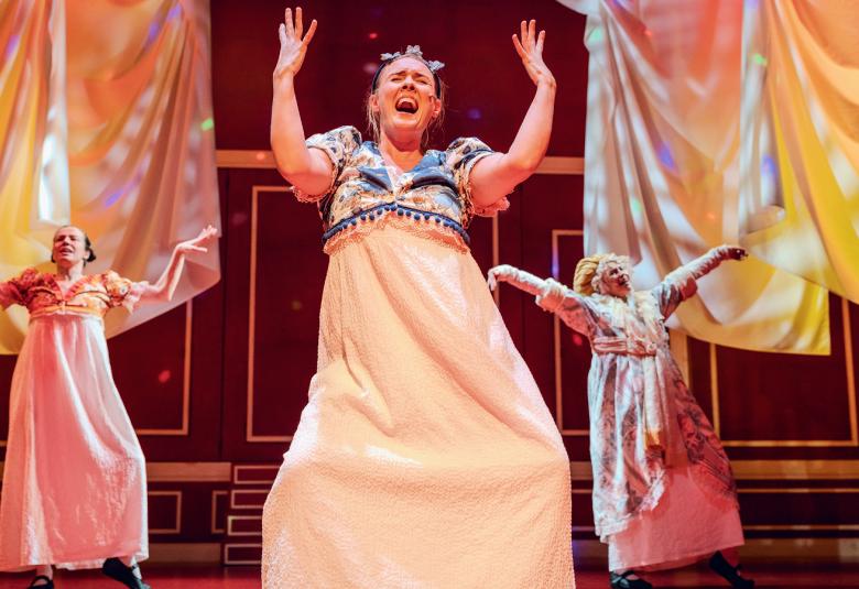 Kuvassa on keskellä nainen pitkässä kellertävässä, pitkässä mekossa ja siinä on sinikuvioinen lyhyt jakku.  Taustalla on molemmin puolin vaaleissa, pitkissä mekoissa tanssimassa. Takana on molemmin puolin roikkumassa keltaista erhoa. Takaseinä on ruskea seinä ja siinä on kultaisin raamein reunustettuja osia.