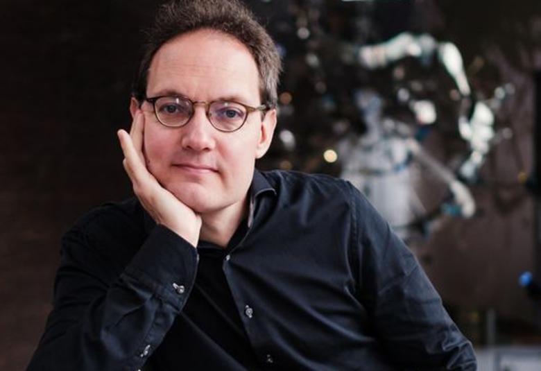 Kuvassa on puolivartalokuvassa Aapo Häkkinen ja hän nojaa oikealla kädellään  poskeensa. Hänellä on musta paita päällään. Tausta on tumma ja sieltä näkyy valoja.