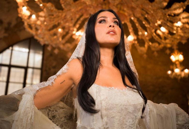 Kuvassa on keskellä Abreu valkoisessa hihattomassa pitsimekossa ja pitsihuntu päässään tummissa, pitkissä hiuksissaan sekä pitää huntua käsivarsillaan. Taustalla on ylhäällä kultainen suuri kattokruunu.