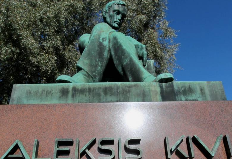 Kuvassa on yläviistoon kuvattu Aleksis Kiven patsas ja ylhäällä istuu Aleksis Kivestä vestetty patsas, jossa hän on istumassa jalat ristissä. Patsas on vihertävä. Alapuolella on ruskeaa graniittia jalustana ja liinä on kohokirjaimin Aleksis Kivi. Taustalla on lehtipuu.