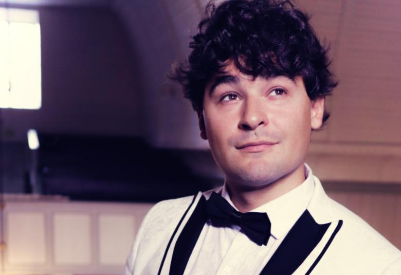 Kuvassa on Amadeus Lundberg puolivartalokuvassa kuvan oikeassa laidassa. Hänellä on valkoinen takki, jossa on kauluksessa mustat kantit. Hänellä on musta rusertti ja valkoinen paita.