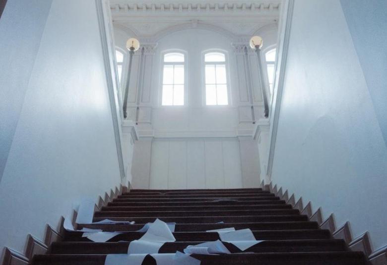 Kuvassa on valkoinen ylöspäin menevä rappukäytävä, jossa on mustilla portailla valkoisia papereita. Ylhäällä on kaksi korkeaa ruutuikkunaa korkealla.