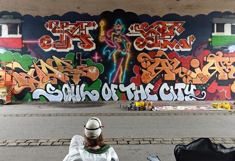 Kuvassa on graffitia taustalla olevalla betoniseinällä sillan alla.  Edessä on katselija valkoisessa kypärässä ja takissa. Välissä on harmaata asfalttia.