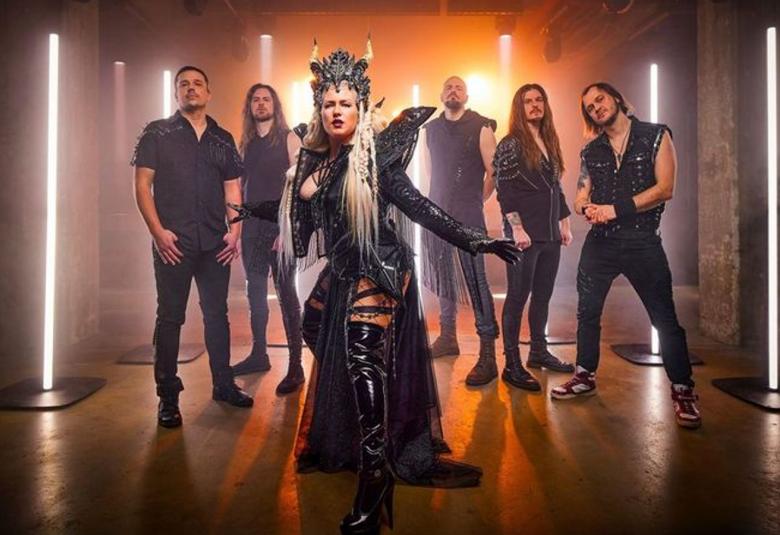 Kuvassa on Battle Beast ja etualalla on naissolisti mustassa näyttävässä puvussa, jossa on korkeat olkapääsiivet ja koristeellinen päähine sarvineen. Taustalla on viisi miest mustissa vaatteissa. Takana pystysuuntaisia led-valoja.