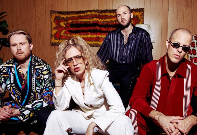 Kuvassa on Haloo Helsingin neljä jäsentä, joista kolme istuu ja yksi seisoo takana. Vasemmalla on mieshenkilökirjavassa takissa ja hänen oikealla puolellaan on nainen valkoisessa housupuvussa. Oikealla istuu mies punaisessa paidassa. Näiden kahden välissä takana seisoo mies, jolla on tumman sininen paita päällään. He ovat ruskeasävyisessä huoneessa. 