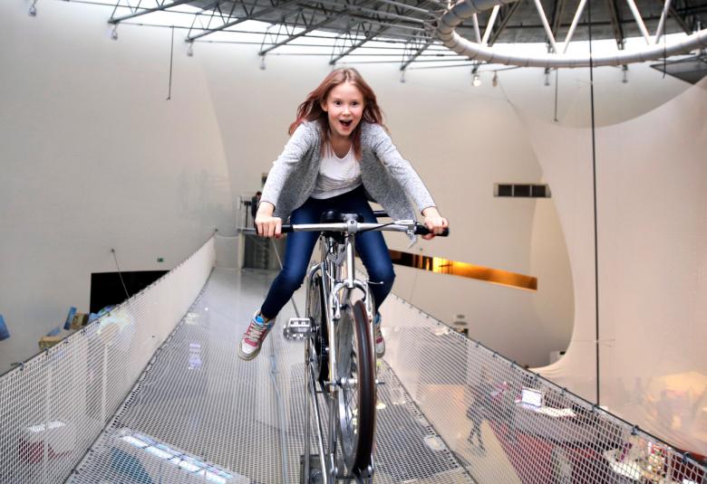 Kuvassa on keskellä nuori tyttö polkupyöräilemässä kiiltävää kourua pitkin metri lattian yläpuolella. Yllä on metallirakenteita. 