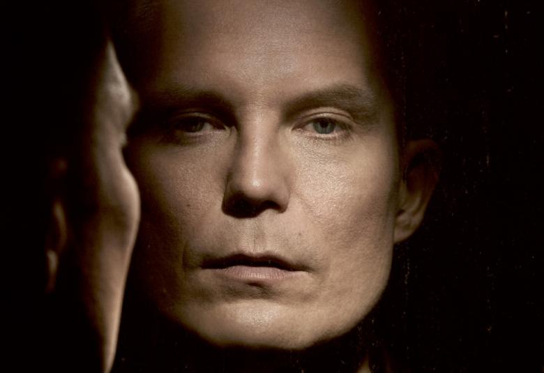 Kuvassa on Jari Tiihosen kasvot ja vasemmalla on sivukuva kasvoista kohti kasvokuvaa. Kuva on musta-valkoinen ja hieman ruskehtava. Tausta on tumma ja kuva on osa kirjan kannesta.